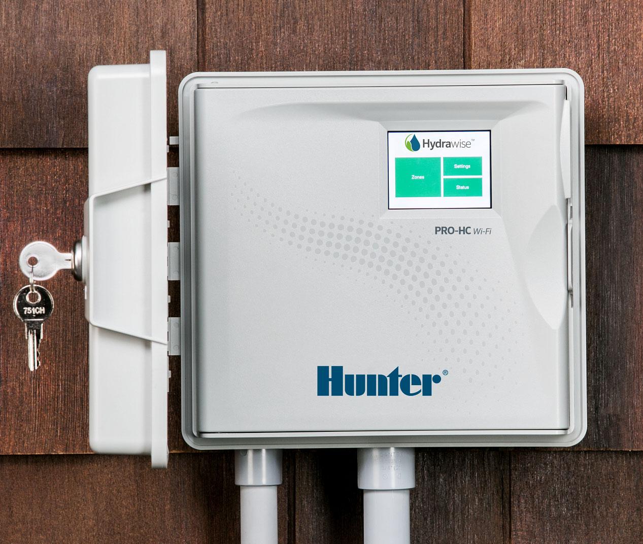 Hunter Hydrawise PRO-HC Wi-Fi sprinkler controller mounted on a home's exterior siding, with its touchscreen showing Zones, Settings, and Status