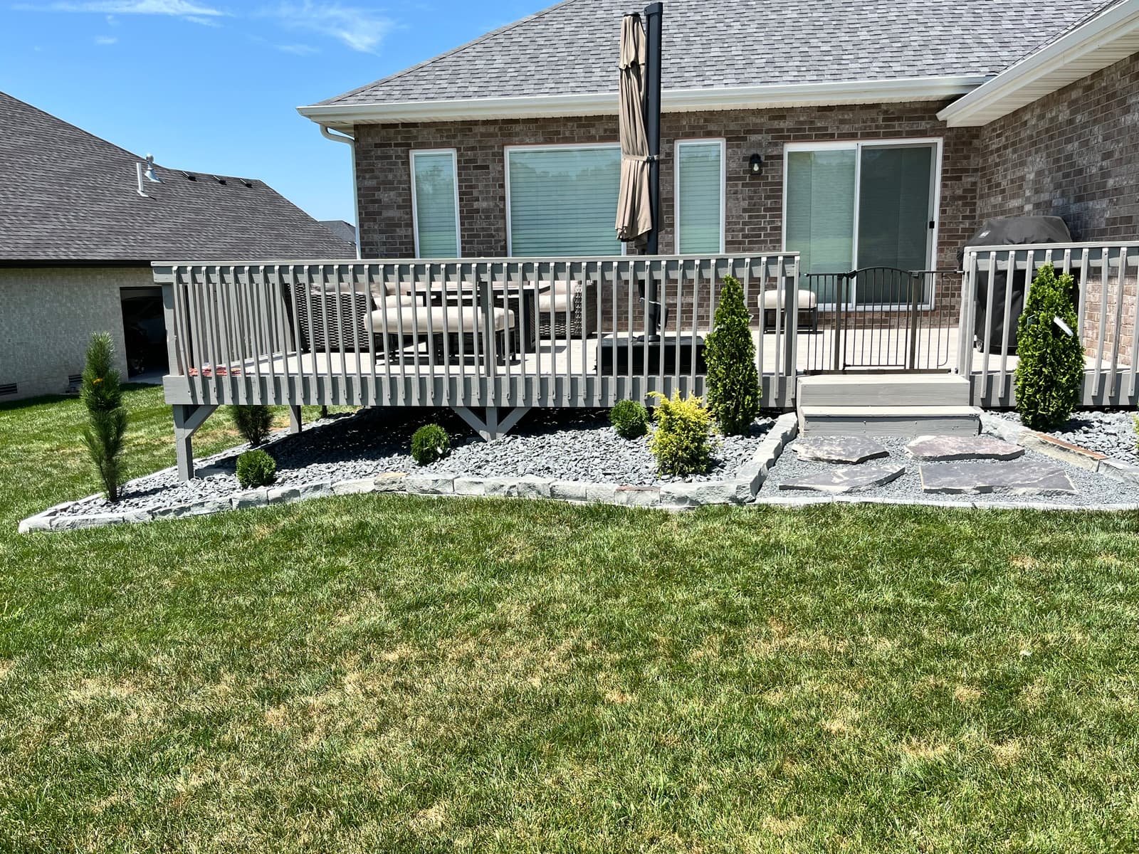 Brick ranch home with a raised deck, fresh white rock beds edged in stone, young evergreens, and a flagstone landing