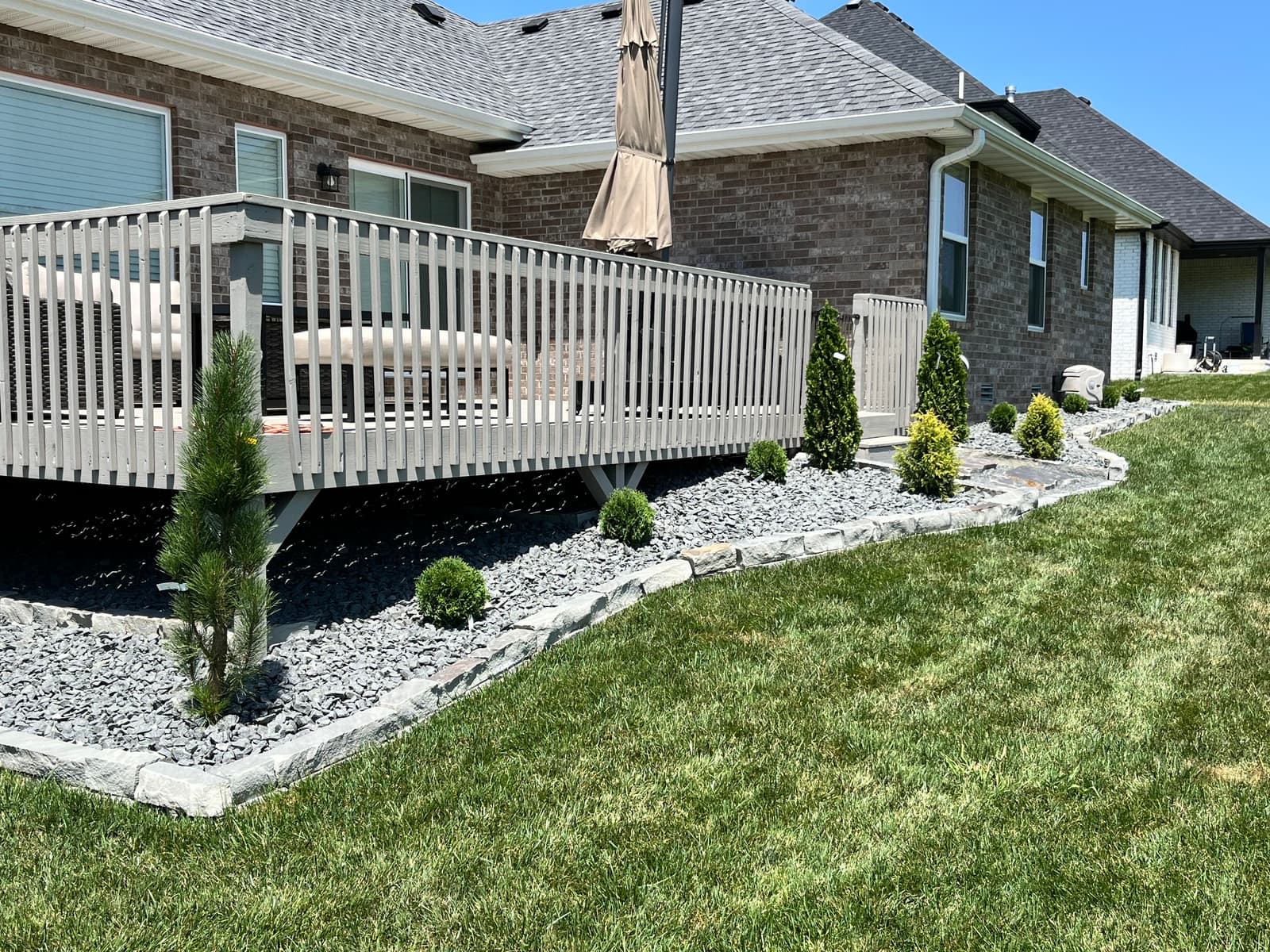 Light rock border wrapping the side of a raised deck with young arborvitae, gold-tipped shrubs, and cut stone edging against the lawn
