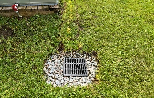 Black square drainage grate set flush in a green lawn and ringed with pale gravel, collecting surface water