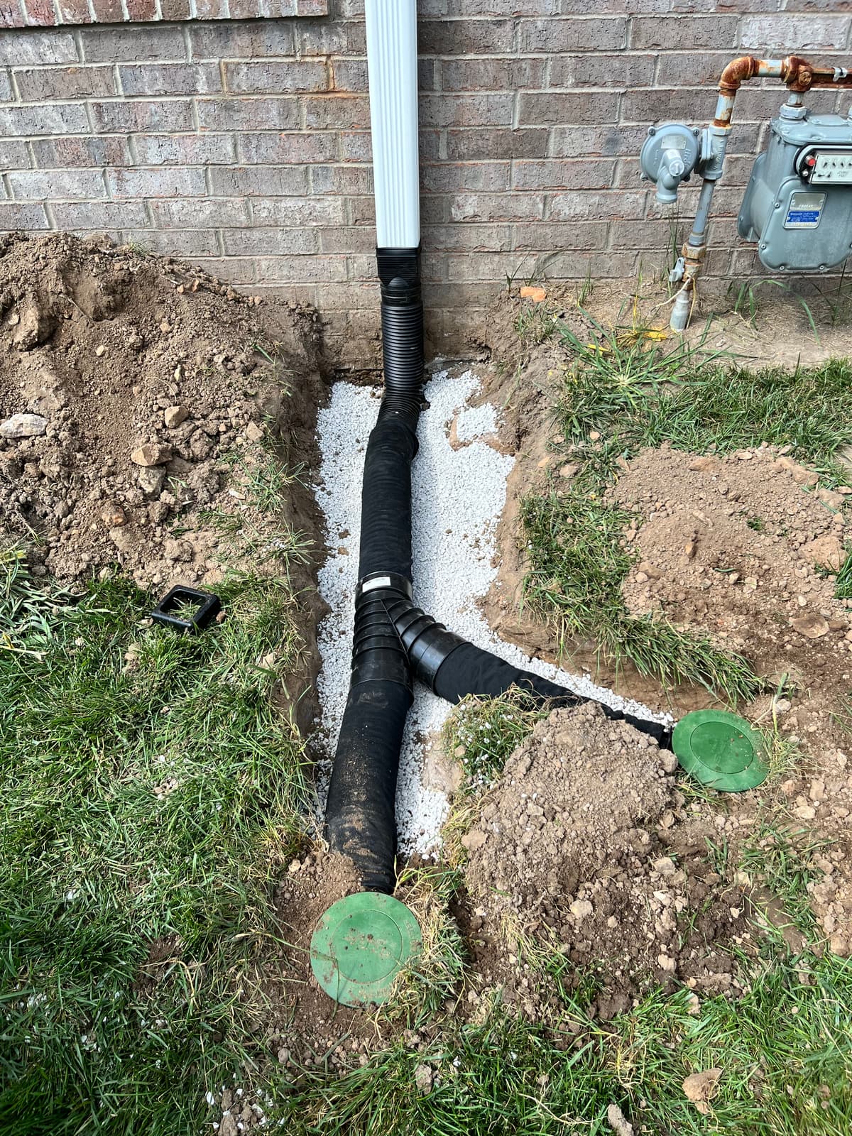 Open trench with corrugated drain pipe bedded in white gravel next to a green irrigation valve box