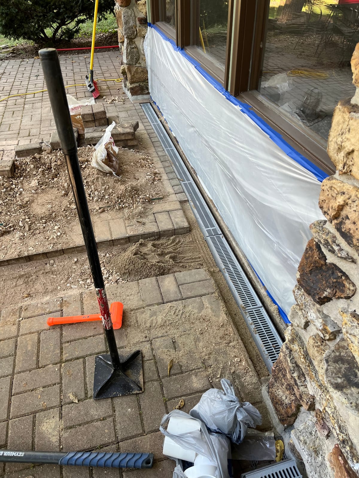 Grated channel drain being set along a stone house foundation with pavers lifted and a hand tamper on the patio