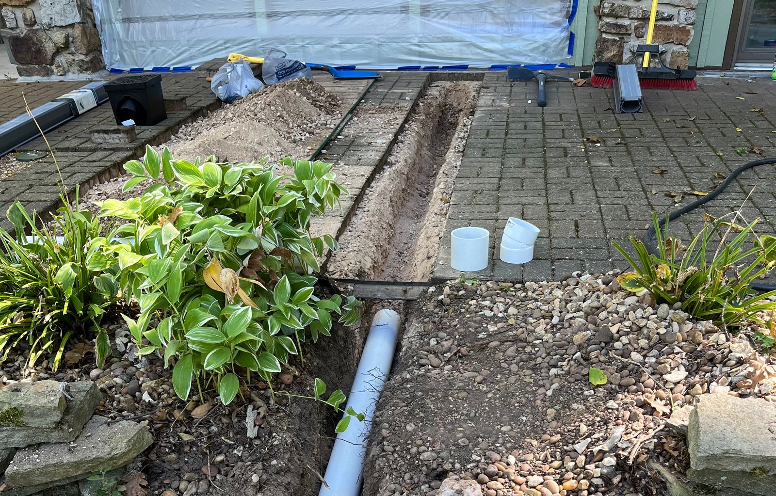 Narrow trench dug through a brick paver patio with PVC fittings and drain parts staged beside it