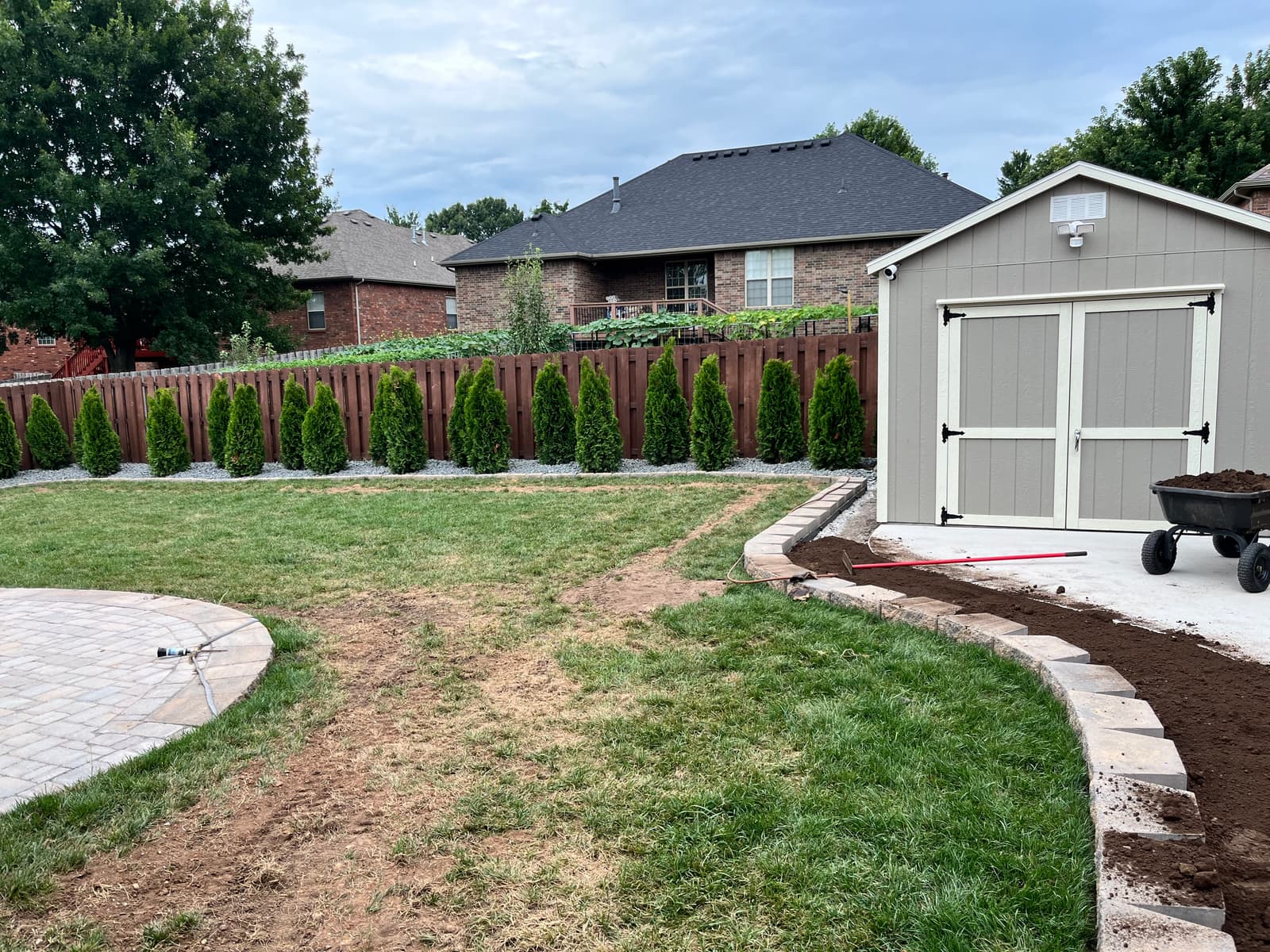 Backyard with a curved paver border running to a storage shed, a paver patio, and a row of young arborvitae along the fence line