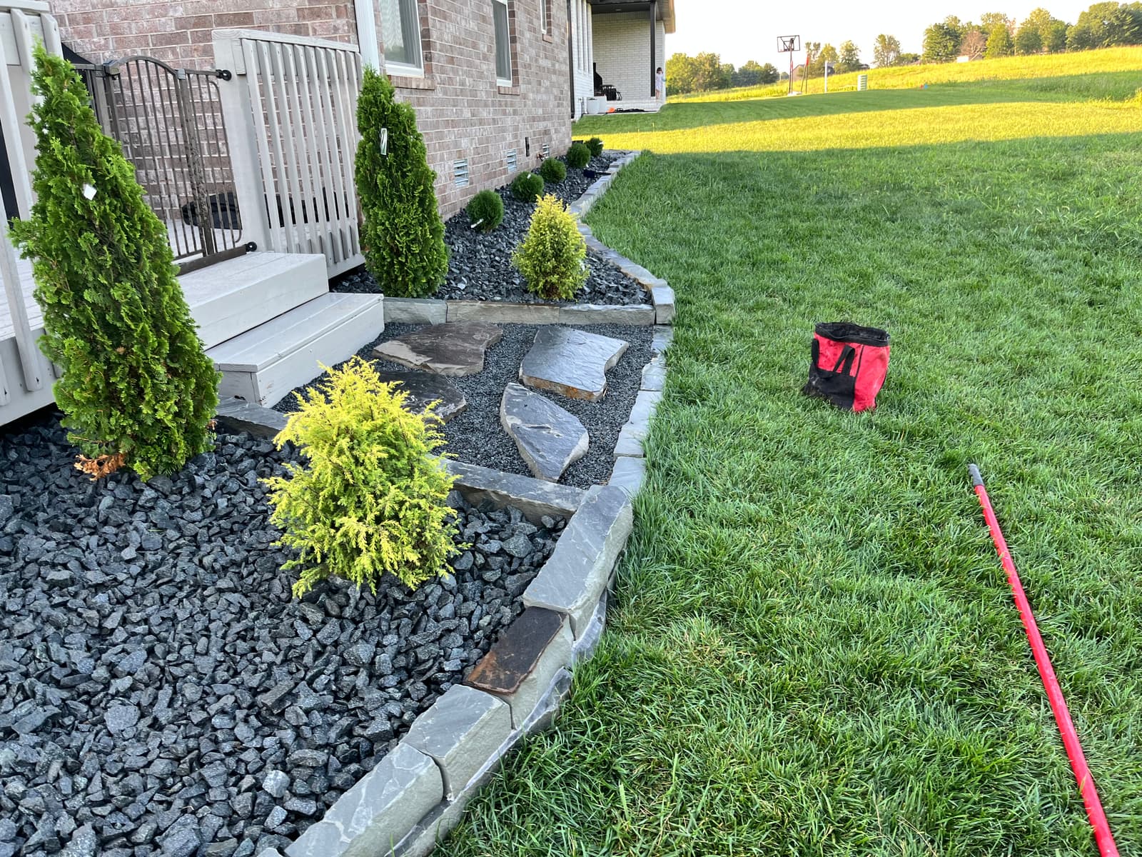 Flagstone stepping stones set through a black rock bed edged in cut stone, leading past arborvitae and gold-tipped shrubs to a porch step
